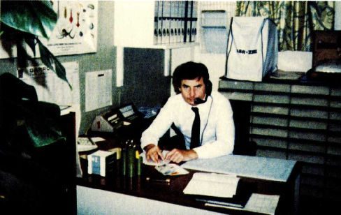A colour photograph of a man in shirtsleeves and tie sitting at an office desk with a telephone, papers and ring binders, filing shelves behind him