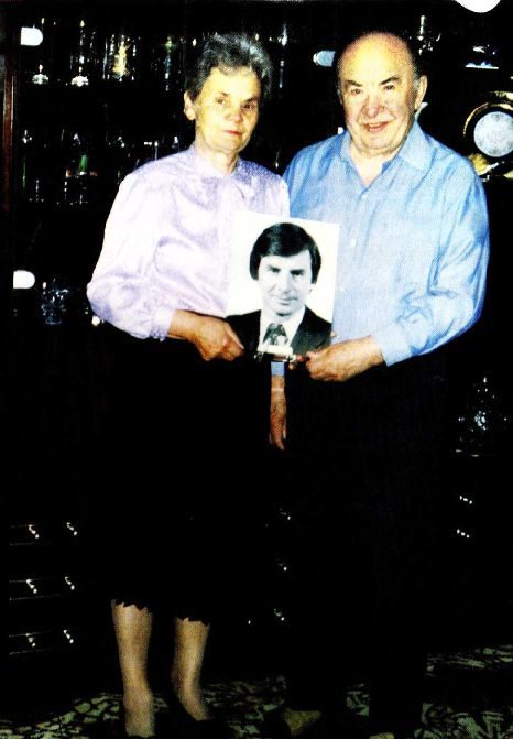 A colour photograph of an elderly woman and an elderly man standing side by side in a dim room, holding a framed portrait photograph of a dark haired man in a suit between them