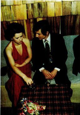 A colour photograph of a young woman in a red evening dress and a young man in a dark suit sitting together on a bench at a dance, glasses and flowers on the table in front of them