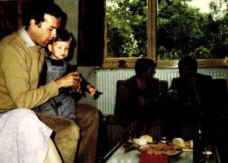 A colour snapshot in a conservatory: a man holding a small child on his lap at a low table, two people seated on a sofa behind them, a garden beyond the windows