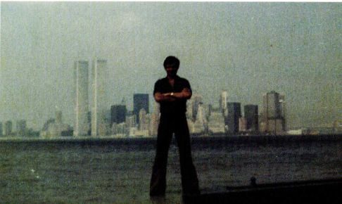 A colour photograph of a man standing at a waterside with his arms folded, the Manhattan skyline and the twin towers of the World Trade Center in haze behind him