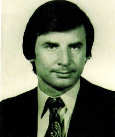 A studio head and shoulders portrait of a dark haired man in a suit and patterned tie, looking at the camera