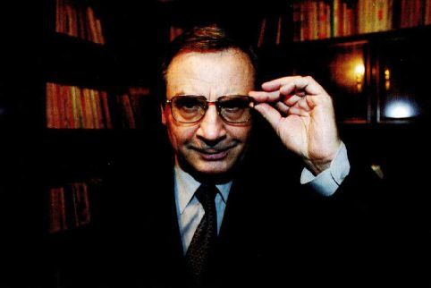 A colour portrait of an older man in a suit and tie in front of full bookshelves, one hand raised to the frame of his glasses