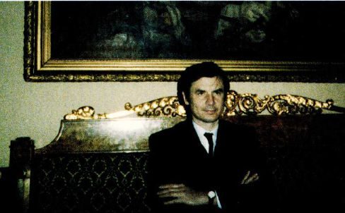 A colour photograph of a man in a dark suit sitting with folded arms on a gilded settee, beneath the lower edge of a large painting in a heavy gilt frame
