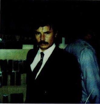 A colour photograph of a man with a moustache in a dark suit and tie standing in front of a display stand of small objects