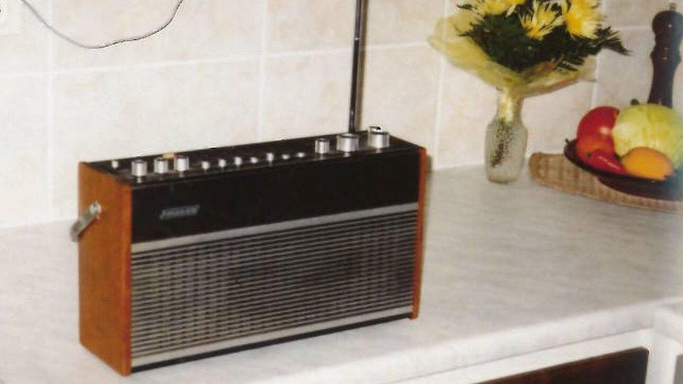 A wood cased portable short-wave receiver standing on a tiled kitchen counter with its telescopic aerial extended, a vase of yellow flowers and a bowl of fruit beside it