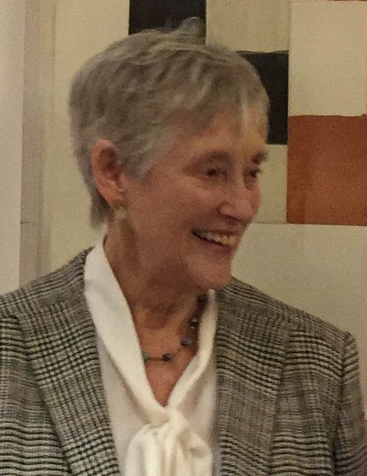 A colour photograph of an older woman with short fair hair speaking, in a dark jacket