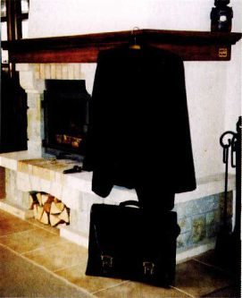 A colour photograph of a dark suit hanging on a beam above a stone fireplace, with a leather briefcase standing on the floor below it