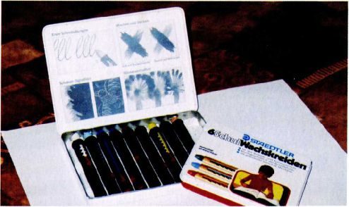 A colour photograph of an open tin of wax crayons with a printed instruction card in the lid, beside a cardboard Staedtler crayon box and a sheet of white paper