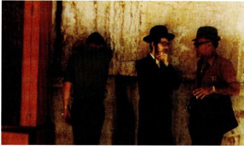A colour photograph of three men in dark coats and hats standing at a large stone wall, one of them with a hand raised to his face