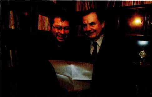 A colour photograph of two men side by side in a book lined room, a younger man in glasses on the left and an older man in a suit on the right, both looking down at a large sheet of paper