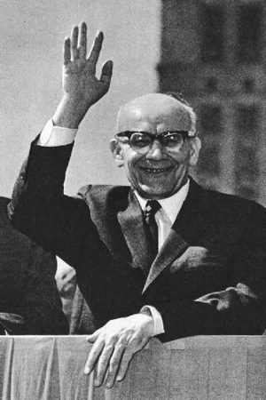 Wladyslaw Gomulka in a dark suit and heavy glasses, waving from a tribune
