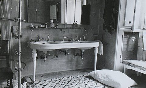 The bathroom of Jan Masaryk's flat on 10 March 1948: a double washbasin on a stand with turned legs, a mirror and bottles on the shelf above it, a shower fitting and a chair at the left, and a pillow lying on the floor beside the patterned mat