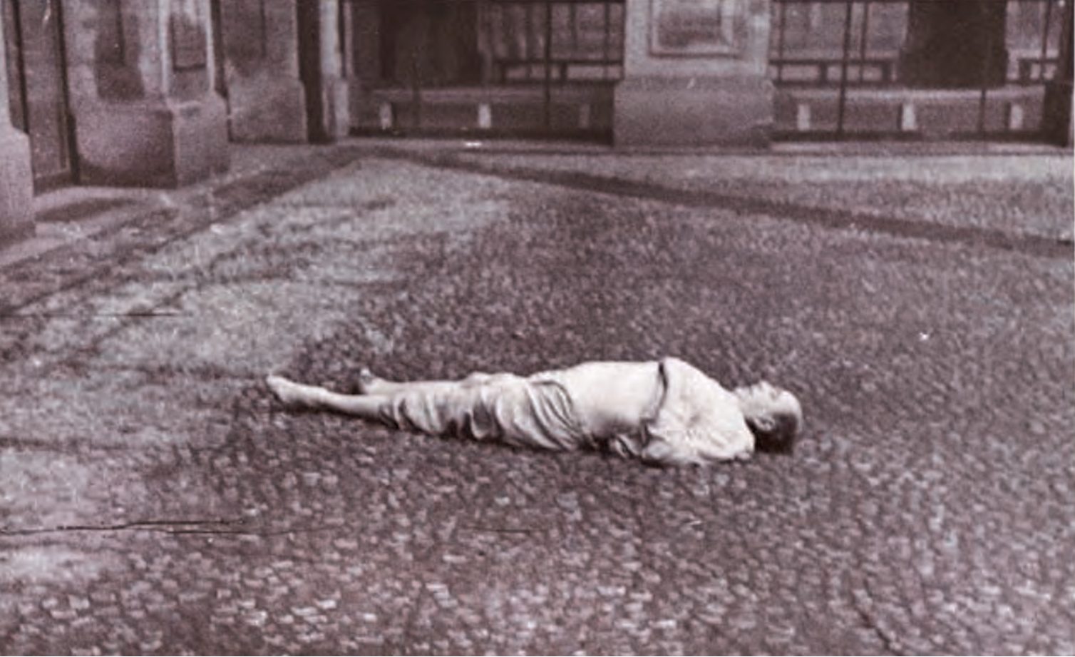 The paved courtyard of the Czernin Palace with the arcade behind: a man in pyjamas lying on his back on the cobbles, barefoot, his head turned to one side