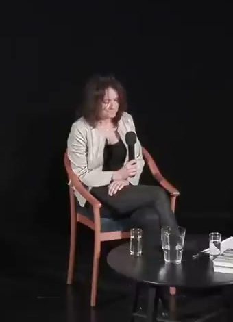 Václava Jandečková seated on a wooden chair on a dark stage holding a microphone, a low table beside her with glasses of water and papers on it