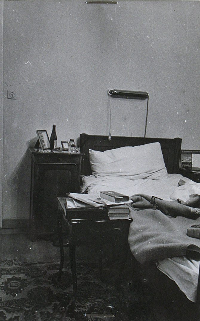 The bedroom of Jan Masaryk's flat on 10 March 1948: the unmade bed with a reading lamp on a bracket above it, a night table carrying a bottle, a glass and a framed picture, and a small table at the bedside piled with books and papers