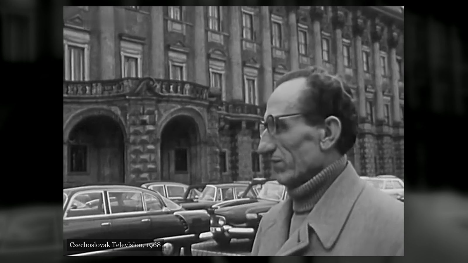 A man in a raincoat and glasses, seen in profile, standing on Loreta Square with the long facade of the Czernin Palace and a row of parked cars behind him, filmed by Czechoslovak Television in 1968