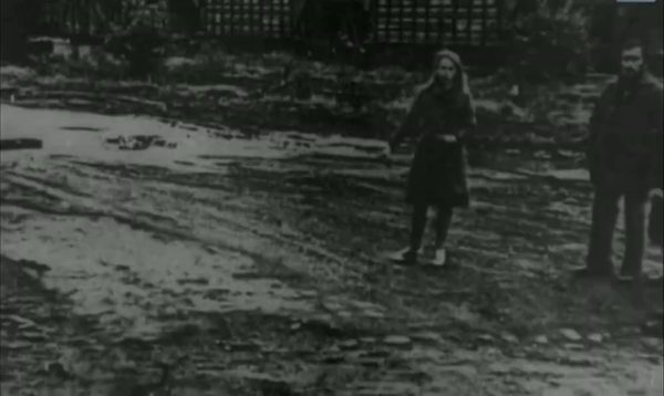 A rutted village track in poor weather: a woman in a headscarf and dark coat standing in the middle of it, a man waiting at the right edge of the frame