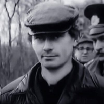 Gennady Mikhasevich in a flat cap and a dark jacket, photographed outdoors among bare trees after his detention, with uniformed officers behind him
