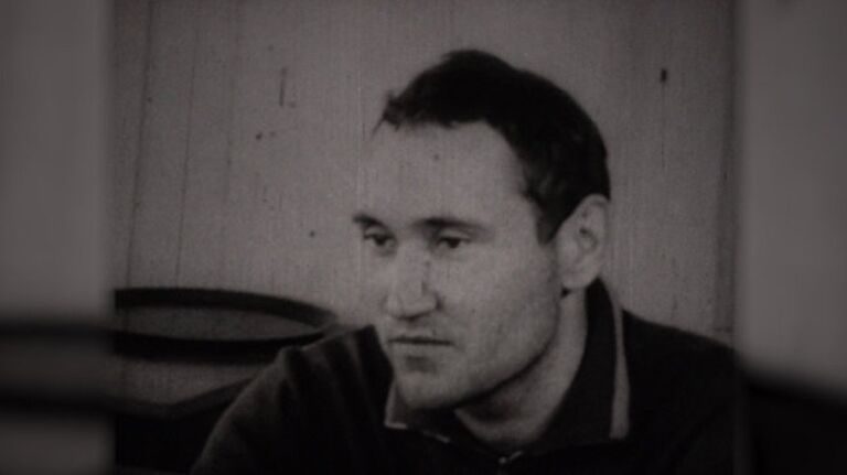 A young man in a dark jacket sitting in a bare room and looking down, photographed during a confrontation with another witness