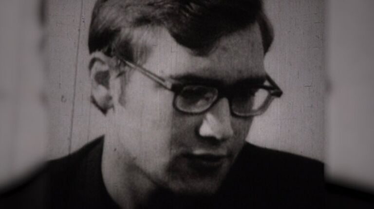 A young man in dark rimmed glasses, head lowered, photographed in a bare room during questioning