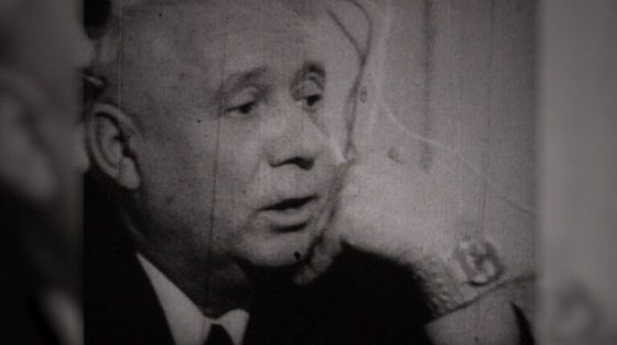 An elderly man in a dark suit and tie, mouth open in mid sentence, filmed close up against a pale wall