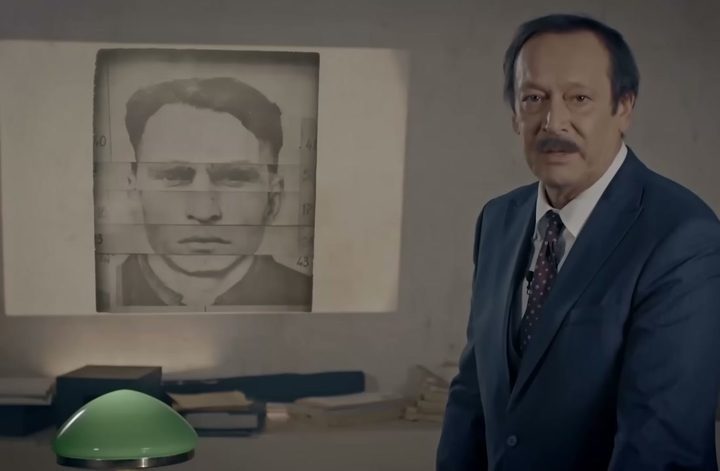 A man's face assembled from horizontal strips of different photographs, forehead, eyes, nose and mouth cut and matched, with measurement figures down both margins, displayed on a wall behind a presenter in a dark suit