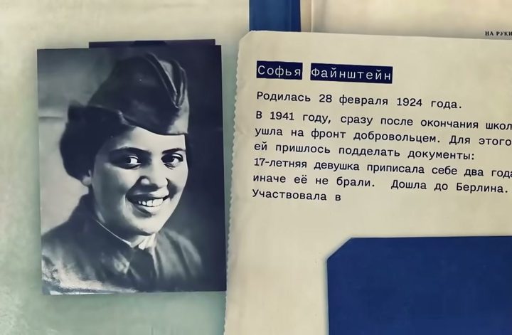 A file page in a documentary: a wartime photograph of a smiling young woman in uniform and forage cap, beside typed Russian lines saying Sofia Fainshtein, born 28 February 1924, left for the front as a volunteer in 1941 after forging her papers to add two years, and reached Berlin