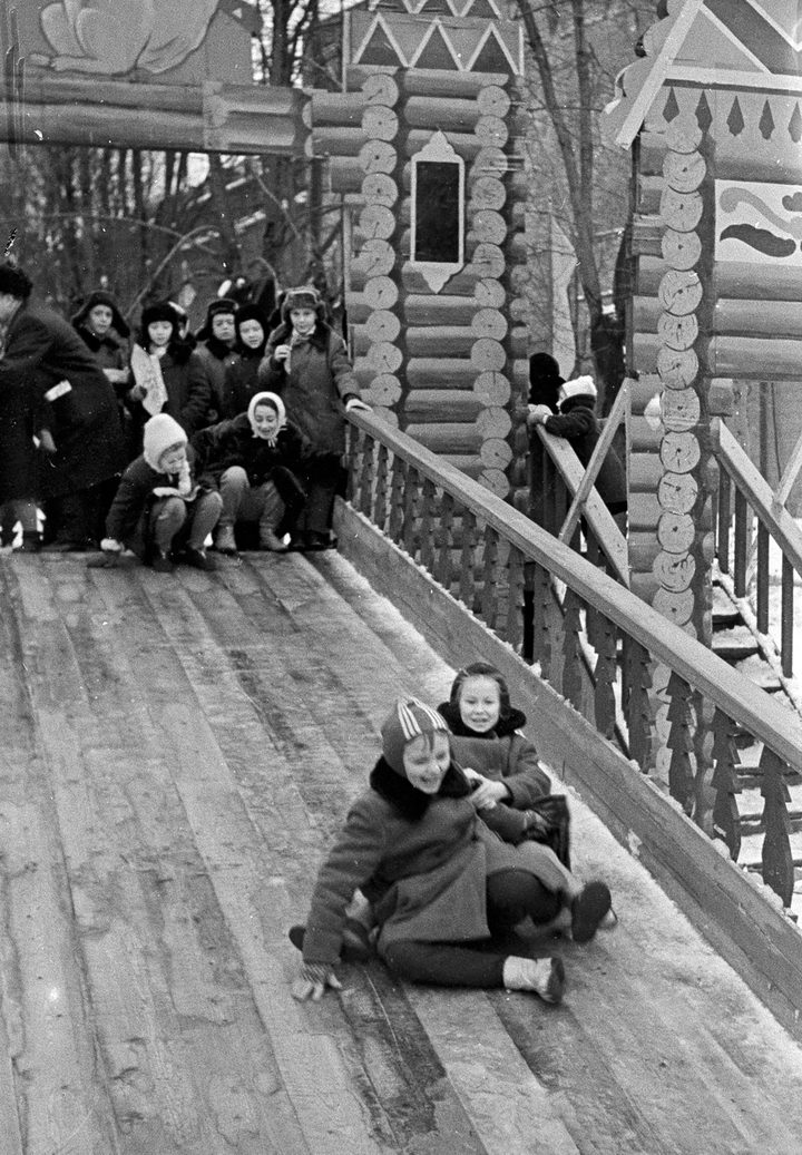 Children in winter coats and fur hats going down a snow slide in the Taynitsky Garden inside the Moscow Kremlin in 1964, adults watching at the side