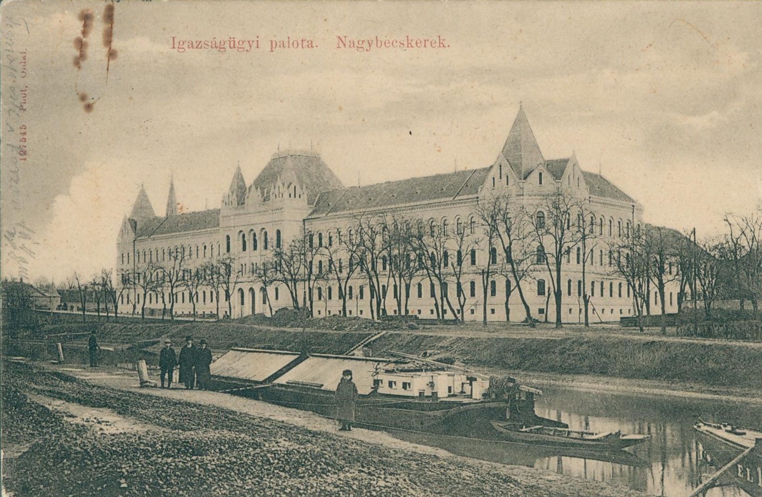 A period postcard captioned Igazsagugyi palota, Nagybecskerek: a long courthouse with corner turrets and rows of arched windows seen across a canal, barges moored at the near bank and four men standing on the towpath