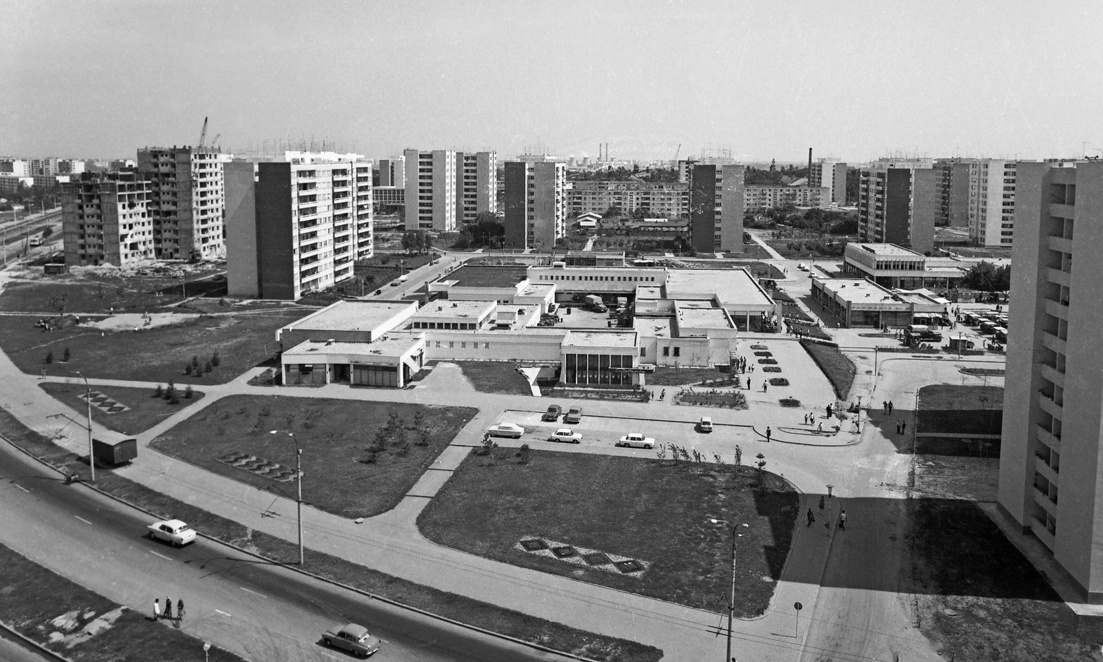 A black and white view over a new Bucharest housing district in 1970, rows of concrete apartment blocks with one still under construction, a low shopping precinct and a plaza with a few parked cars