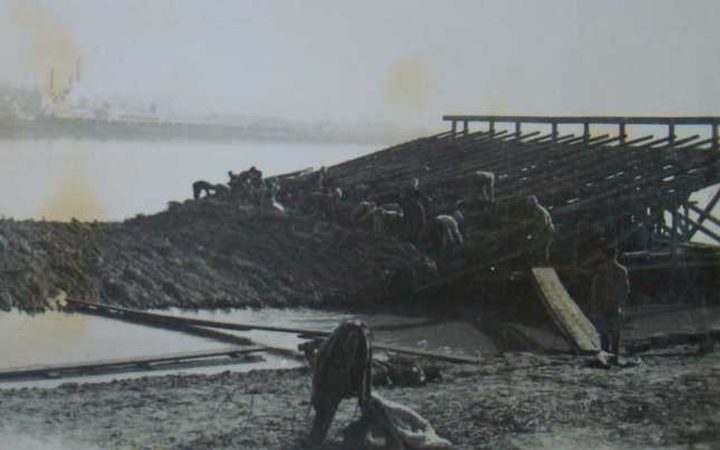 Men working by hand on a timber and earth embankment running into the water, a guard standing at the near end, industrial buildings on the far bank