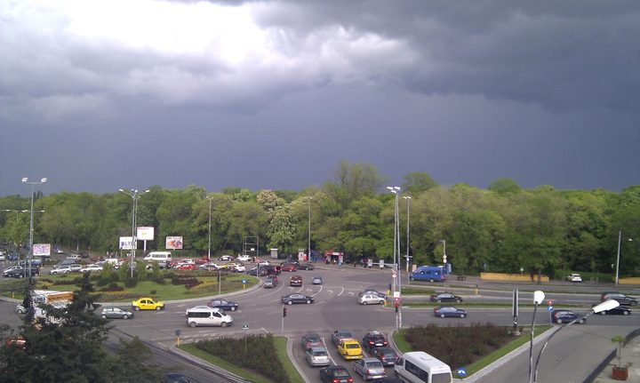 The edge of Herastrau Park in Bucharest seen from above under a storm sky: a boulevard junction with traffic in the foreground and the park's dense tree line running the width of the picture behind it