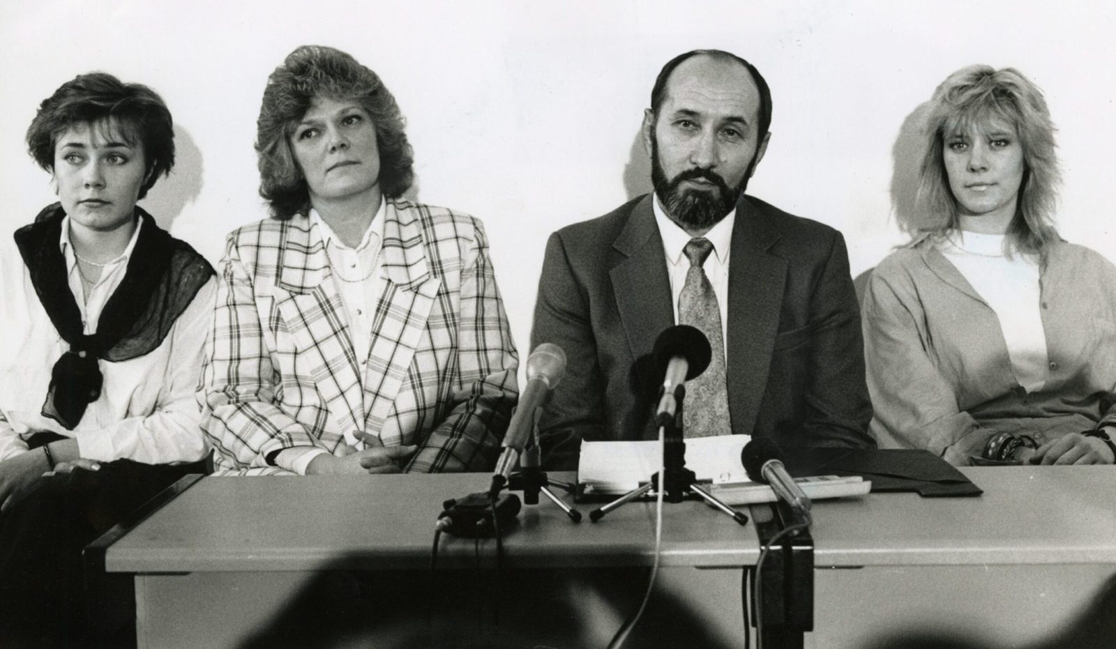 A black and white photograph of Nikola Stedul at a table of microphones with his wife Shirley beside him and their two daughters at either end of the table