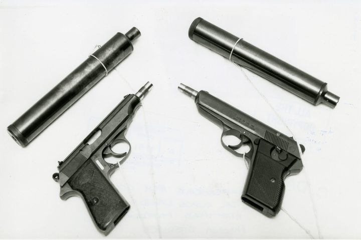 A black and white photograph of two small automatic pistols with threaded muzzles and two long cylindrical silencers laid out on a white surface, each piece carrying a tied exhibit label; the slide of the right hand pistol is stamped vzor 70 cal 765 made in Czechoslovakia