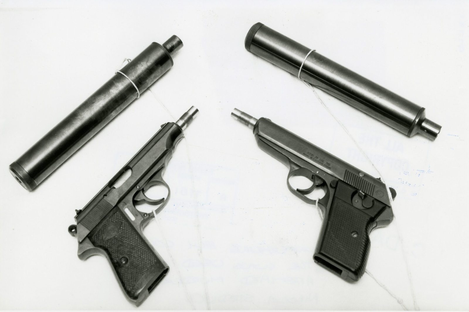 A black and white photograph of two small automatic pistols with threaded muzzles and two long cylindrical silencers laid out on a white surface, each piece carrying a tied exhibit label; the slide of the right hand pistol is stamped vzor 70 cal 765 made in Czechoslovakia