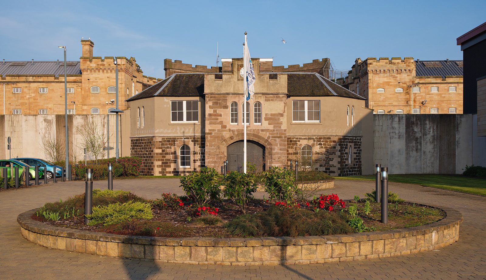 The entrance of Perth prison: a sandstone gatehouse with a castellated tower over an arched gateway, flanked by the battlemented walls and barred windows of the old prison blocks behind a high perimeter wall
