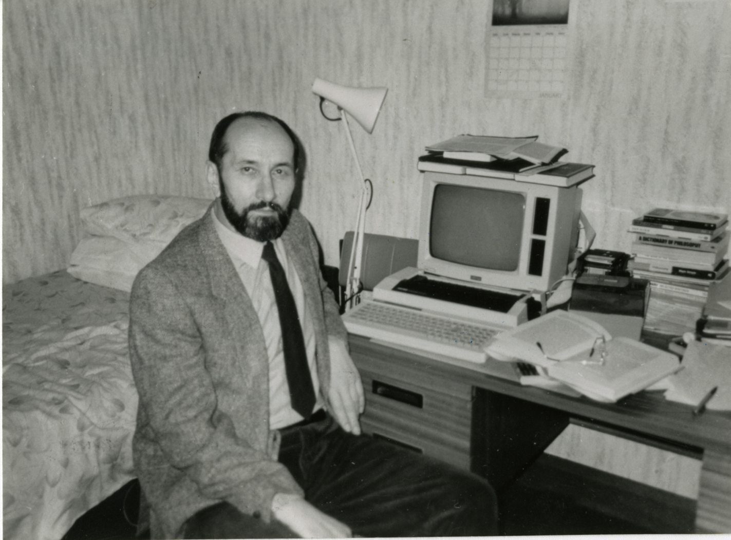 A black and white photograph of Nikola Stedul, bearded and in a jacket and tie, sitting on the edge of a bed beside a desk with an early home computer, an anglepoise lamp, papers and a stack of books