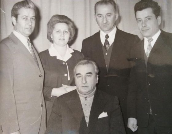 A black and white group photograph in front of a curtain: four people standing, three men in suits and a woman, and in the centre, seated, an older man in a dark suit with a pocket square, Dumitru Ceacanica