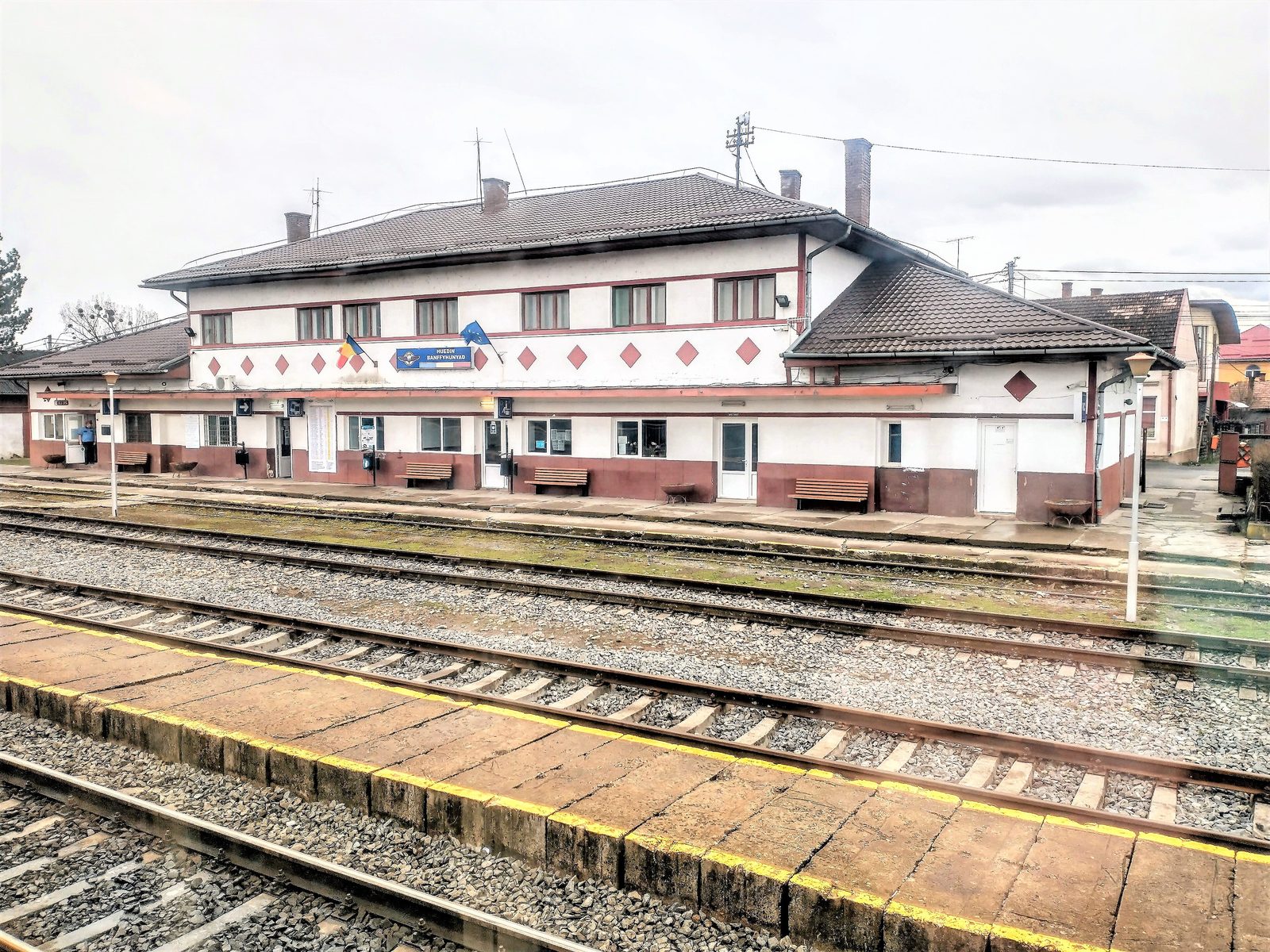 A colour photograph of the platform side of Huedin railway station, a long white building with dark red trim and diamond decoration, carrying a blue sign that reads HUEDIN BANFFYHUNYAD, with benches under the canopy and three tracks in the foreground