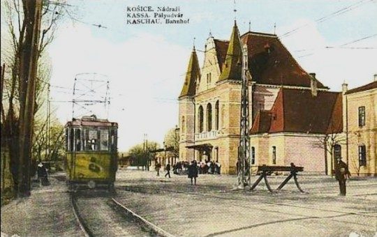 A coloured postcard of the railway station at Košice with a tram in the street outside, captioned in Czech, Hungarian and German