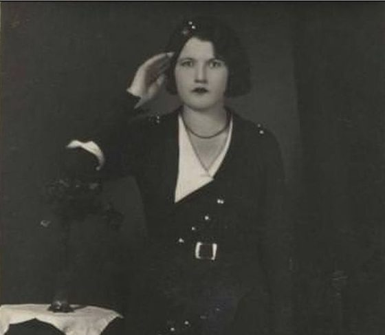 A young woman in a dark belted dress standing beside a table with flowers, in a studio portrait of the early 1930s