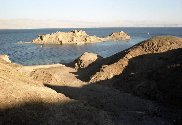 A small rocky island with the ruins of a fortress, off a sandy shore and low brown hills