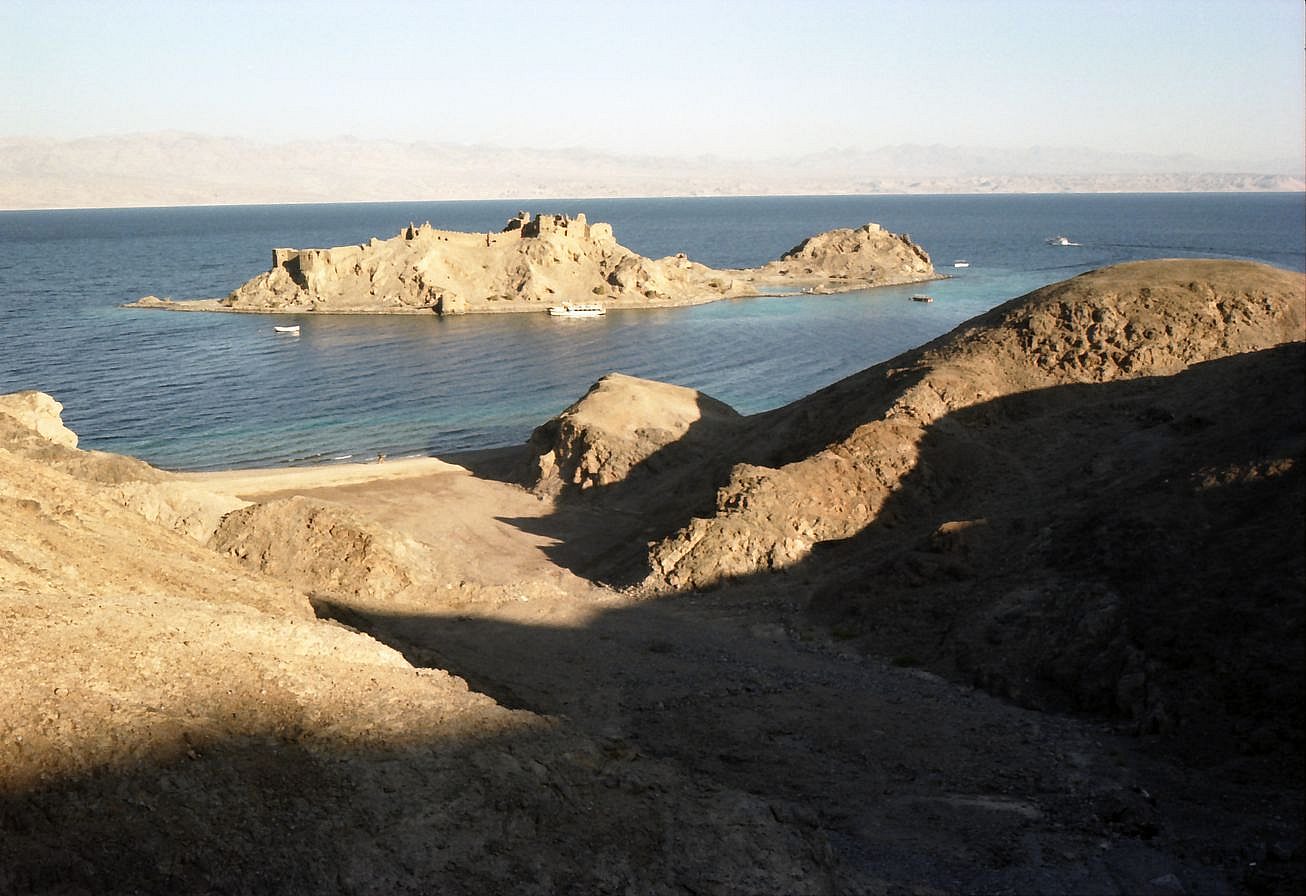 A small rocky island with the ruins of a fortress, off a sandy shore and low brown hills