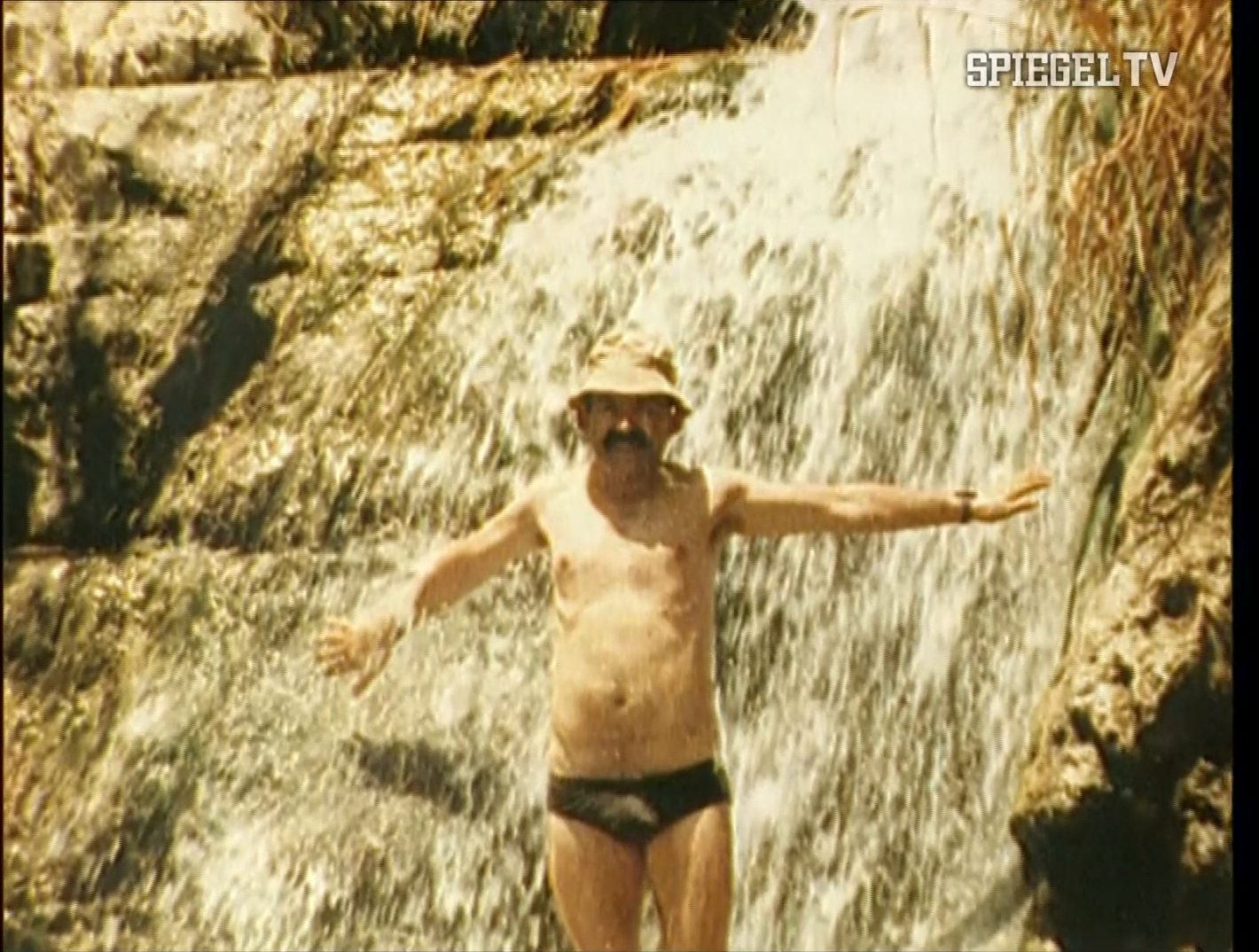 A moustached man in swimming trunks and a hat standing under a small waterfall