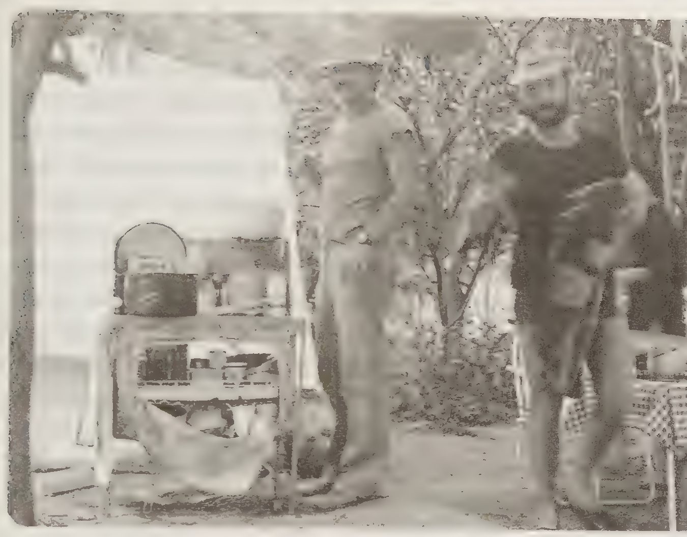A grainy black and white photograph of two men standing outdoors beside a table with cooking things