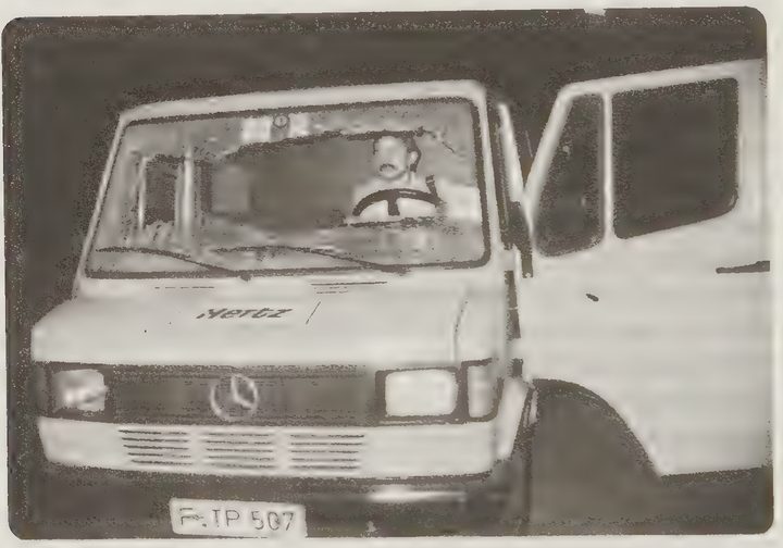 A black and white photograph of the front of a van with a broken windscreen and a moustached man sitting at the wheel