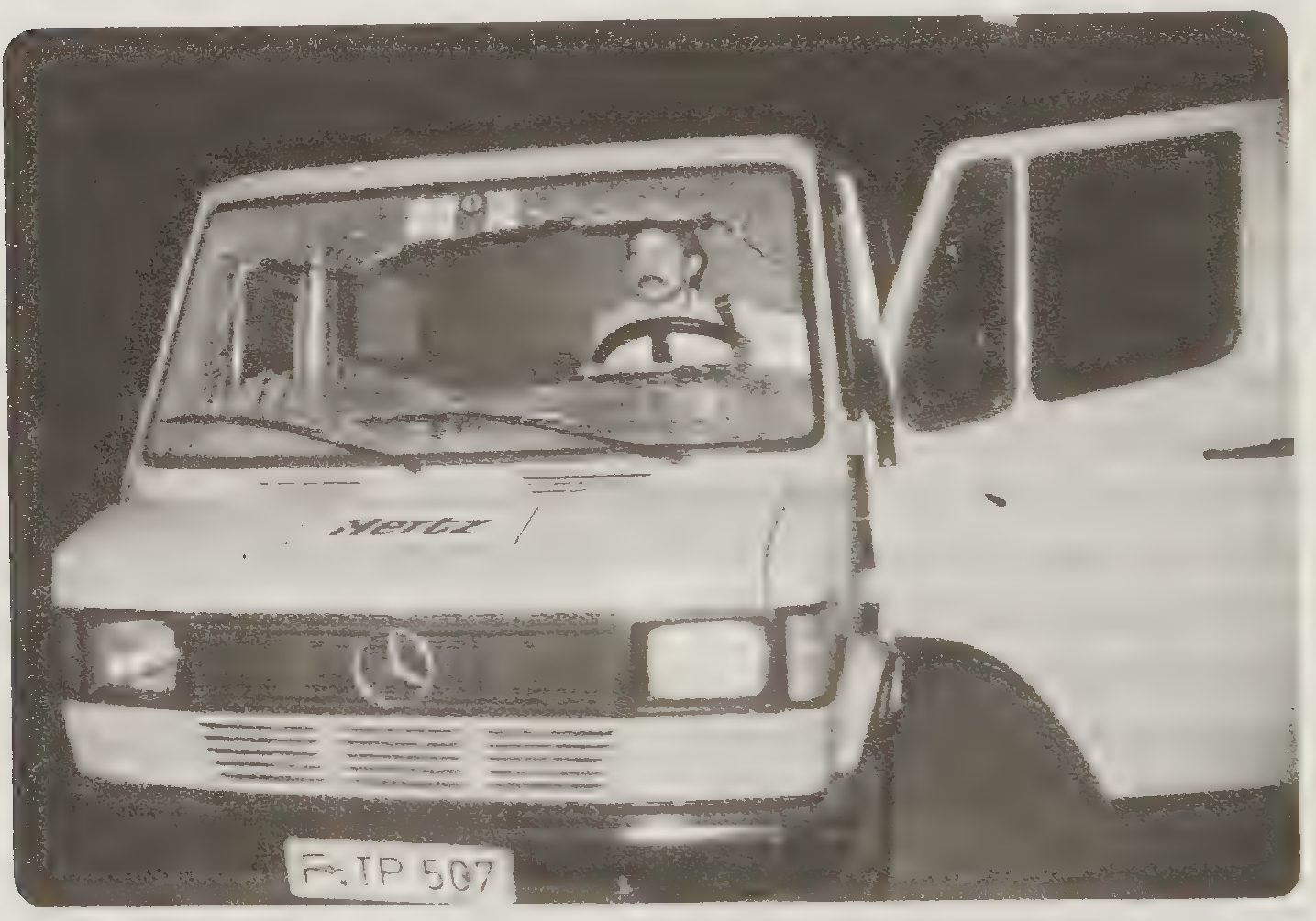 A black and white photograph of the front of a van with a broken windscreen and a moustached man sitting at the wheel
