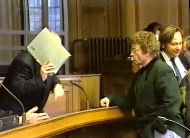 In a wood-panelled courtroom a man hides his face behind a pale green folder while a curly-haired man in a green jacket stands at the rail