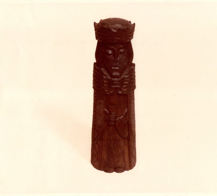 A tall carved figure of dark stained wood, crowned and robed, holding a small cup in both hands, photographed standing on cream photographic paper